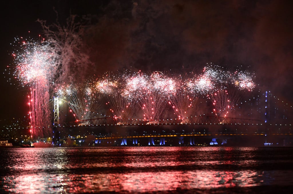 Fogos de artifício iluminam o céu noturno sobre uma ponte, com reflexos visíveis na superfície da água em Florianópolis, um dos melhores lugares para curtir o Réveillon no Brasil.