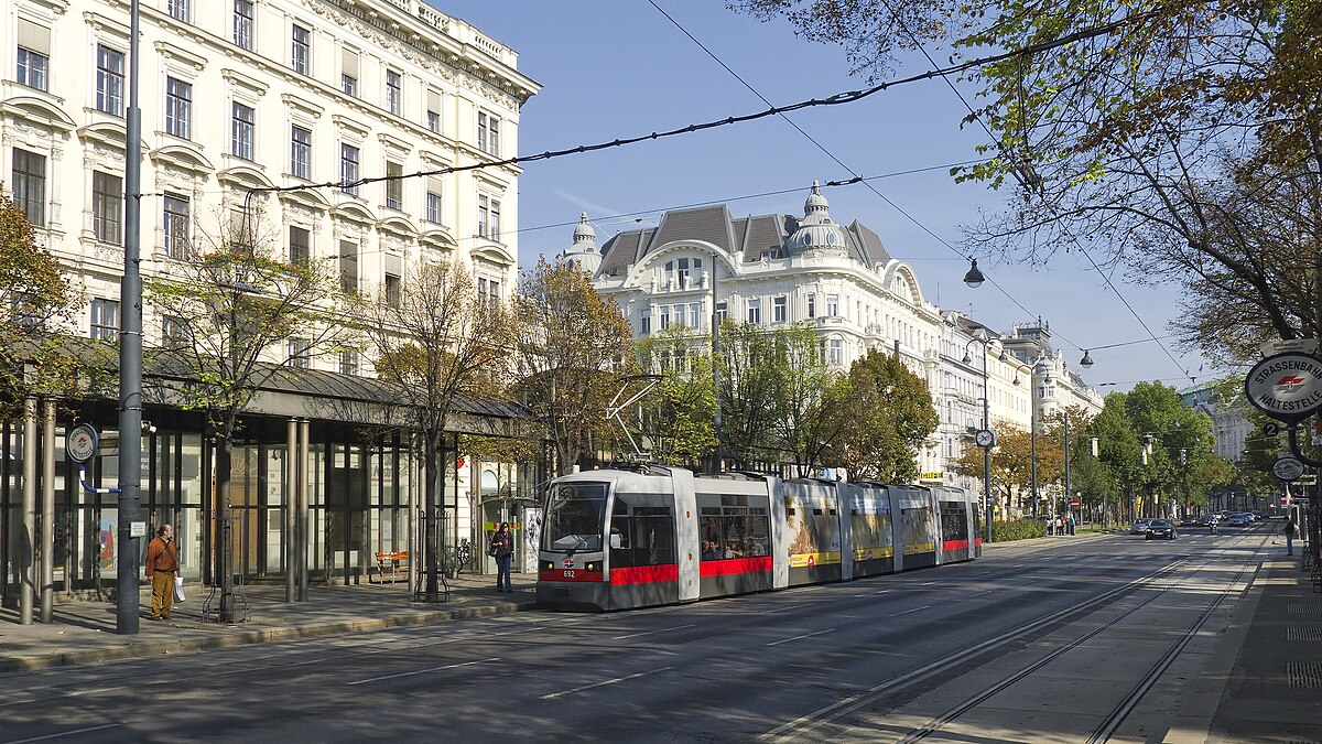Um bonde vermelho e branco viaja por uma rua na Ringstrasse. Ao longo da rua há edifícios históricos sob um céu claro.