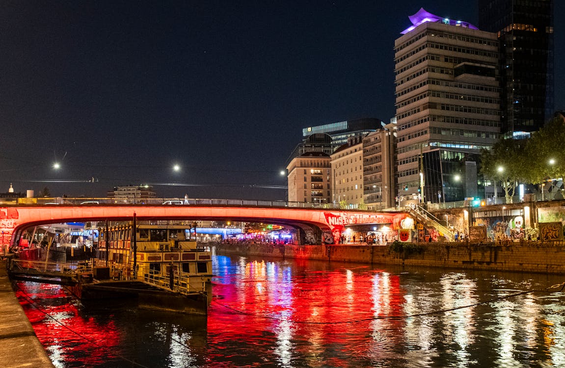 Vista noturna do canal de Danúbio com pontes e prédios iluminados. Reflexos de luzes vermelhas e brancas brilham na água. A foto ilustra o post de o que fazer em Viena.