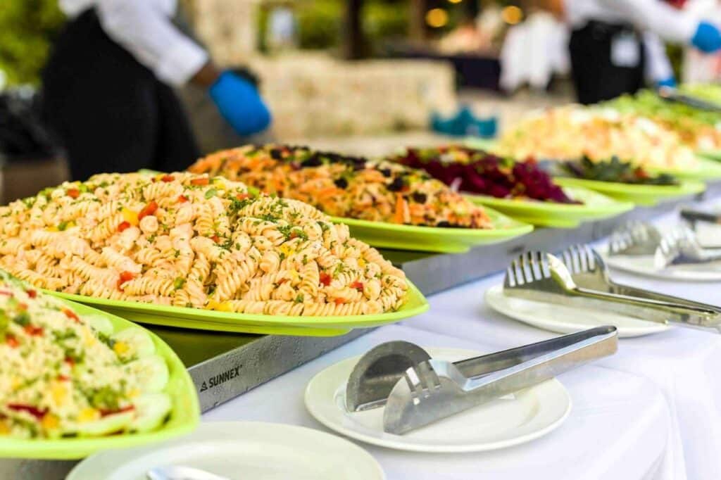 Um serviço de buffet no Sonesta Maho Beach Resort com bandejas de salada de macarrão sobre uma mesa com talheres e pratos, com uma pessoa usando luvas azuis por perto.