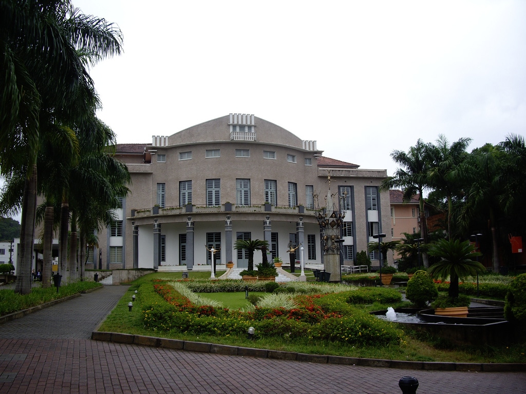 Teatro Carlos Gomes com janelas em arco, cercado por palmeiras e um jardim paisagístico com uma pequena fonte na frente.