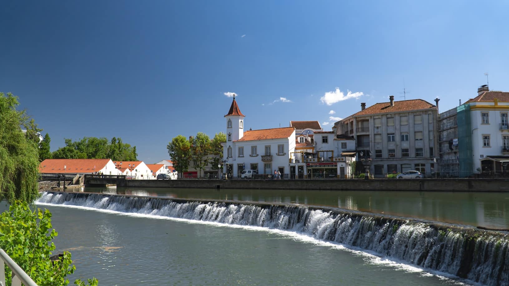 Vista do rio mostrando uma cachoeira, edifícios brancos com telhados vermelhos, uma torre do relógio e um céu azul claro ao fundo, na cidade de Tomar, uma opção de roteiro para Portugal