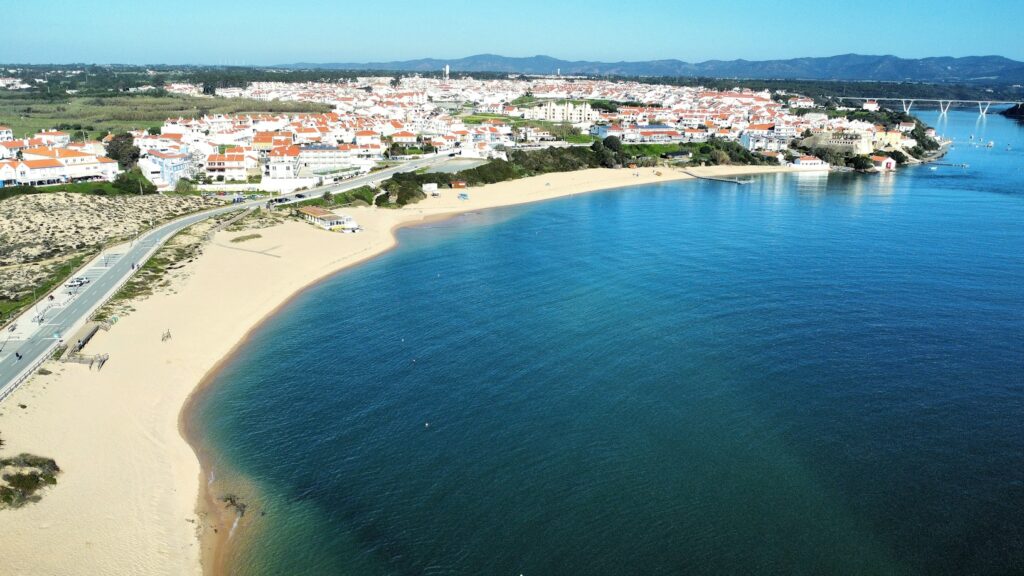 Vista aérea de uma cidade costeira com edifícios brancos e telhados vermelhos, uma praia de areia dourada e um mar azul calmo sob um céu limpo.
