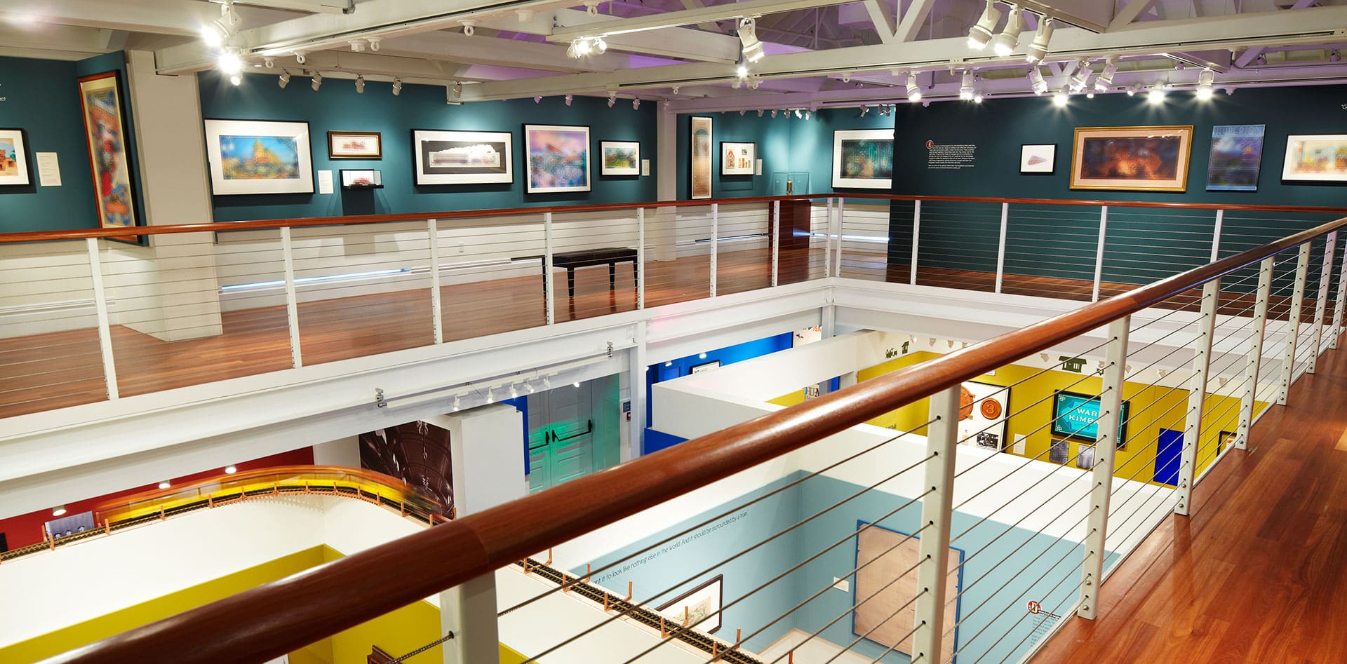 Walt Disney Family Museum de uma galeria de arte moderna com obras de arte emolduradas em paredes azul-petróleo, piso de madeira e uma varanda superior com corrimão de metal. Imagem para ilustrar post sobre museus em San Francisco.