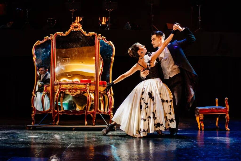 Um casal executa uma dança dramática em trajes formais no palco, com um espelho ornamentado e um banco ao fundo.