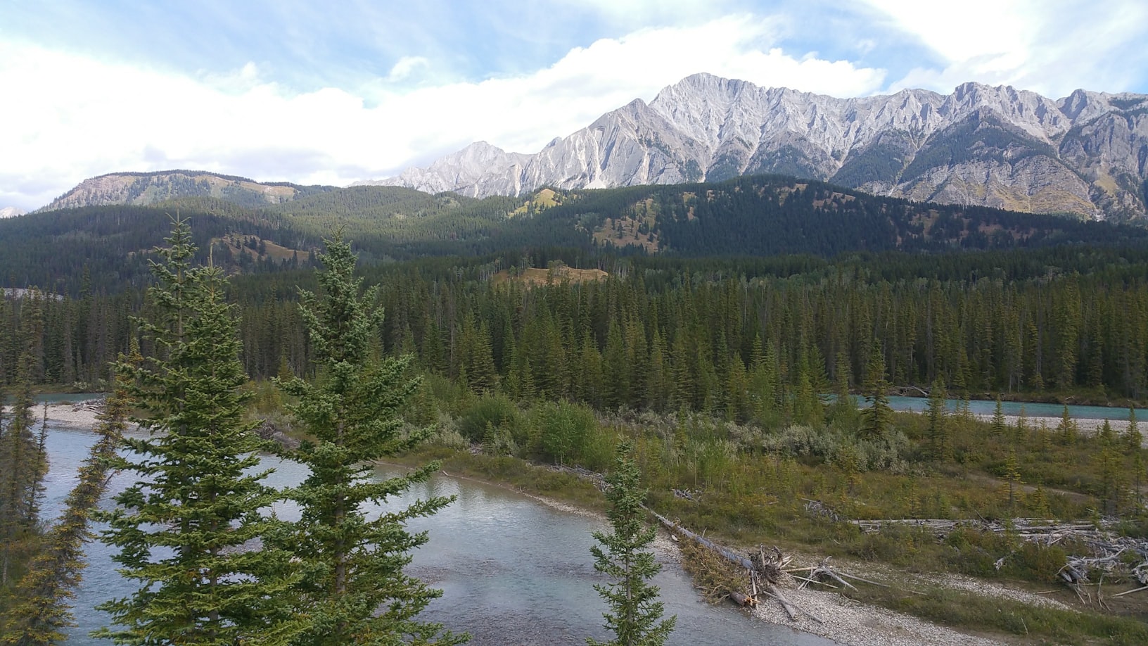 Uma paisagem cênica com um rio em primeiro plano, pinheiros e montanhas ao fundo, sob um céu parcialmente nublado em Banff National Park, Alberta, no Canadá