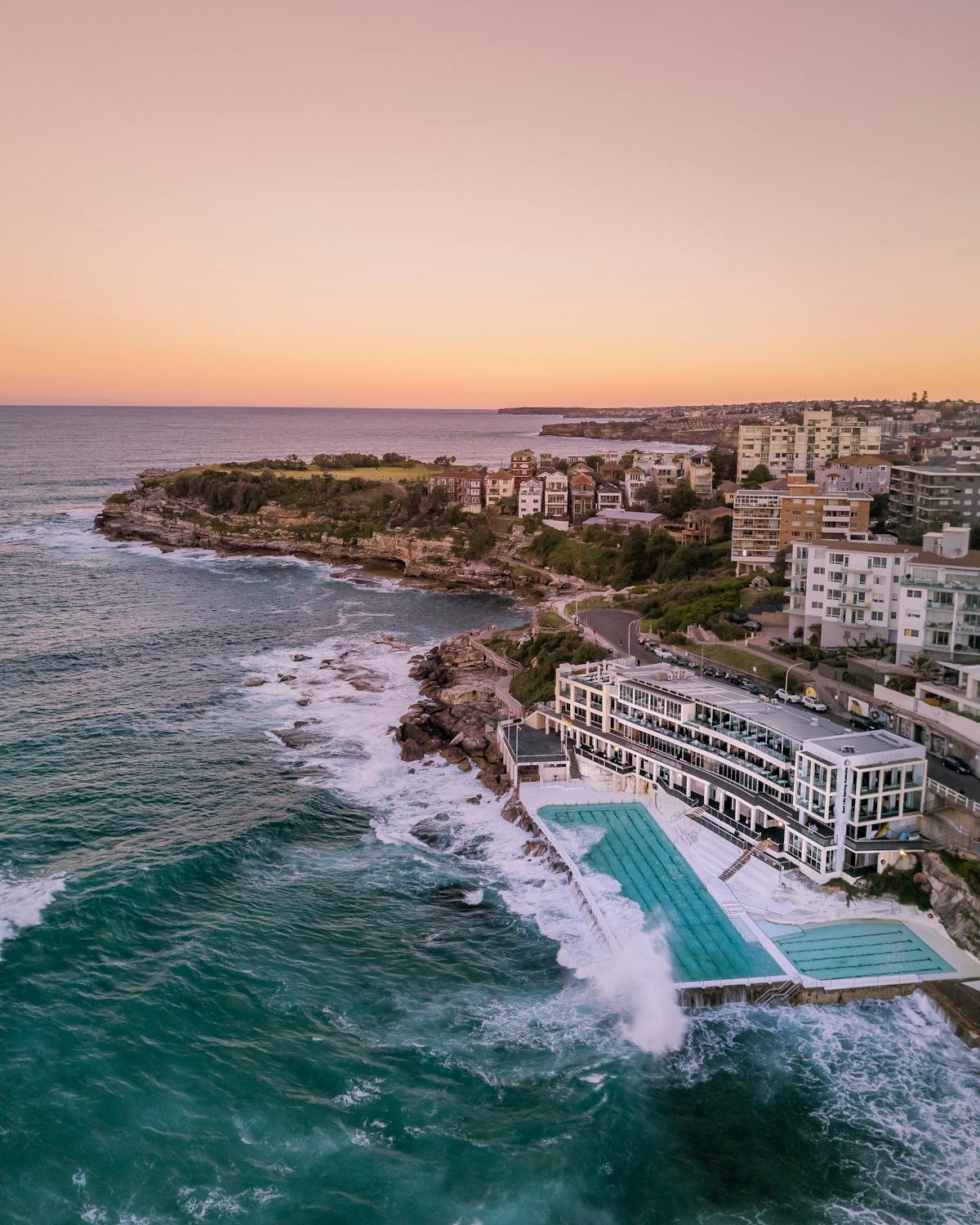 Vista aérea de uma piscina à beira-mar na Bondi Beach e edifícios perto de um litoral rochoso ao pôr do sol, com ondas quebrando contra a praia. A foto está na lista de o que fazer em Sydney.