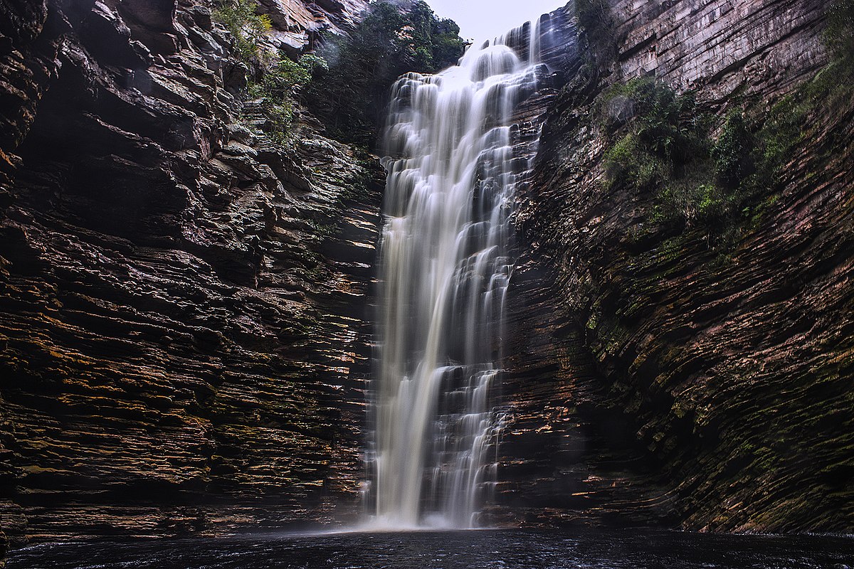 Cachoeira do Buracão desce em cascata por um penhasco rochoso até uma piscina abaixo, cercada por paredões de pedra e vegetação esparsa. A foto ilustra o post de o que fazer na Chapada Diamantina.