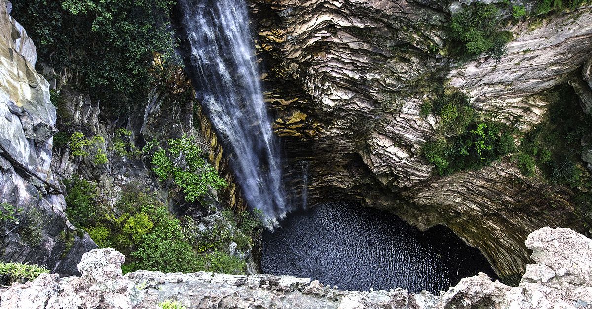A Cachoeira do Buracão desce em cascata até uma bacia rochosa cercada por vegetação exuberante e penhascos escarpados, vista de um ponto alto.