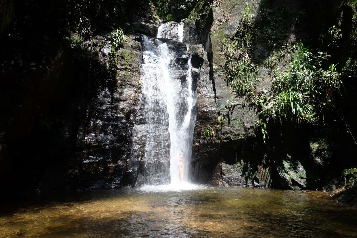 Uma cachoeira desce por uma parede rochosa até formar uma piscina transparente cercada por vegetação exuberante em um dia ensolarado. Essa é a paisagem de uma das trilhas no Rio de Janeiro.