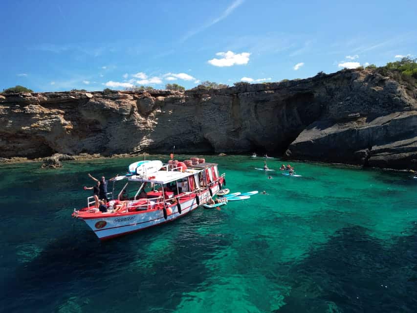 Um barco vermelho e branco ancorado em águas azul-turquesa perto de penhascos rochosos, com pessoas praticando stand up paddle nas proximidades sob um céu azul.
