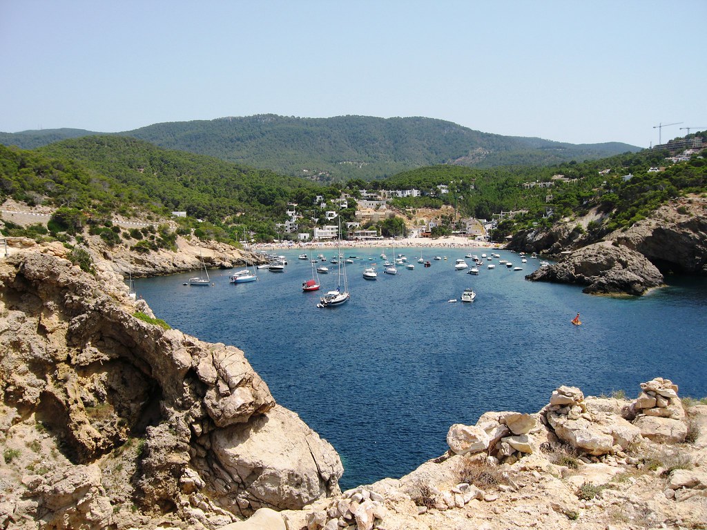 Um litoral rochoso com vista para uma baía calma repleta de barcos ancorados, cercada por colinas verdes e uma pequena cidade litorânea. Essa é a Cala Vadella, uma das praias em Ibiza recomendadas.