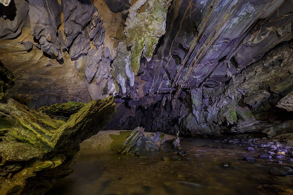 O interior de uma caverna mal iluminada, com formações rochosas irregulares e um pequeno riacho correndo pelo chão rochoso. Essa é a Caverna Água Suja, acessada por uma das trilhas em SP.