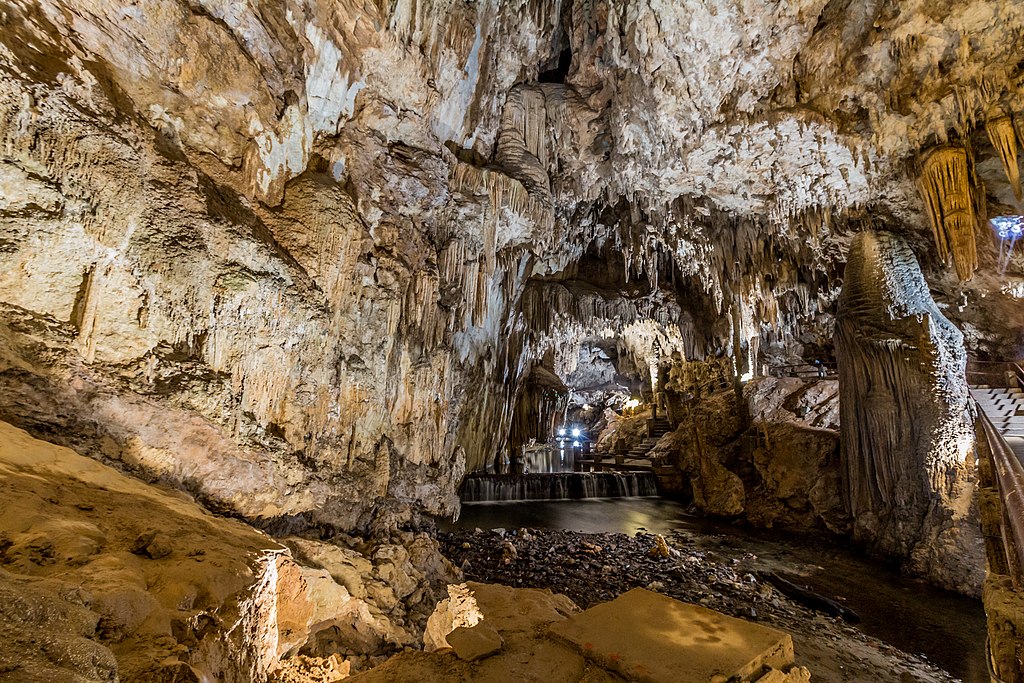 Interior de uma grande caverna com estalactites e estalagmites, iluminada por luzes artificiais, com uma piscina de água no chão. Essa é a Caverna do Diabo, acessada por uma das trilhas em SP.