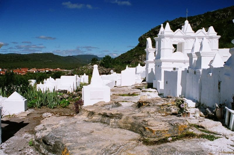 Cemitério Bizantino em Mucugê com mausoléus e tumbas brancas em uma colina rochosa sob um céu azul claro, com vegetação e colinas distantes ao fundo. A foto ilustra o post de o que fazer na Chapada Diamantina.