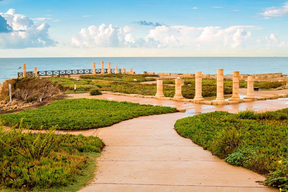 Antigas colunas da Cesareia Marítima de pedra ficam perto de um caminho costeiro com vegetação, com vista para um mar calmo sob um céu parcialmente nublado. Ilustram o que fazer em Israel.
