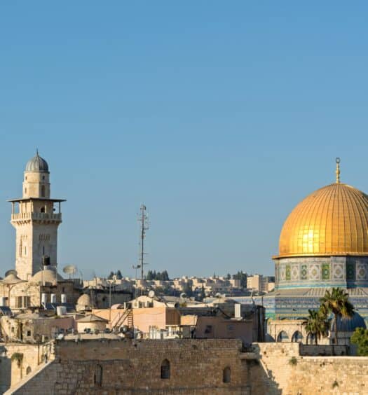 Vista do Domo da Rocha com sua cúpula dourada, cercado por outros edifícios e um alto minarete sob um céu azul claro em Jerusalém.
