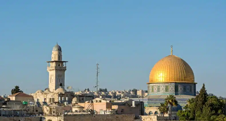Vista do Domo da Rocha com sua cúpula dourada, cercado por outros edifícios e um alto minarete sob um céu azul claro em Jerusalém.