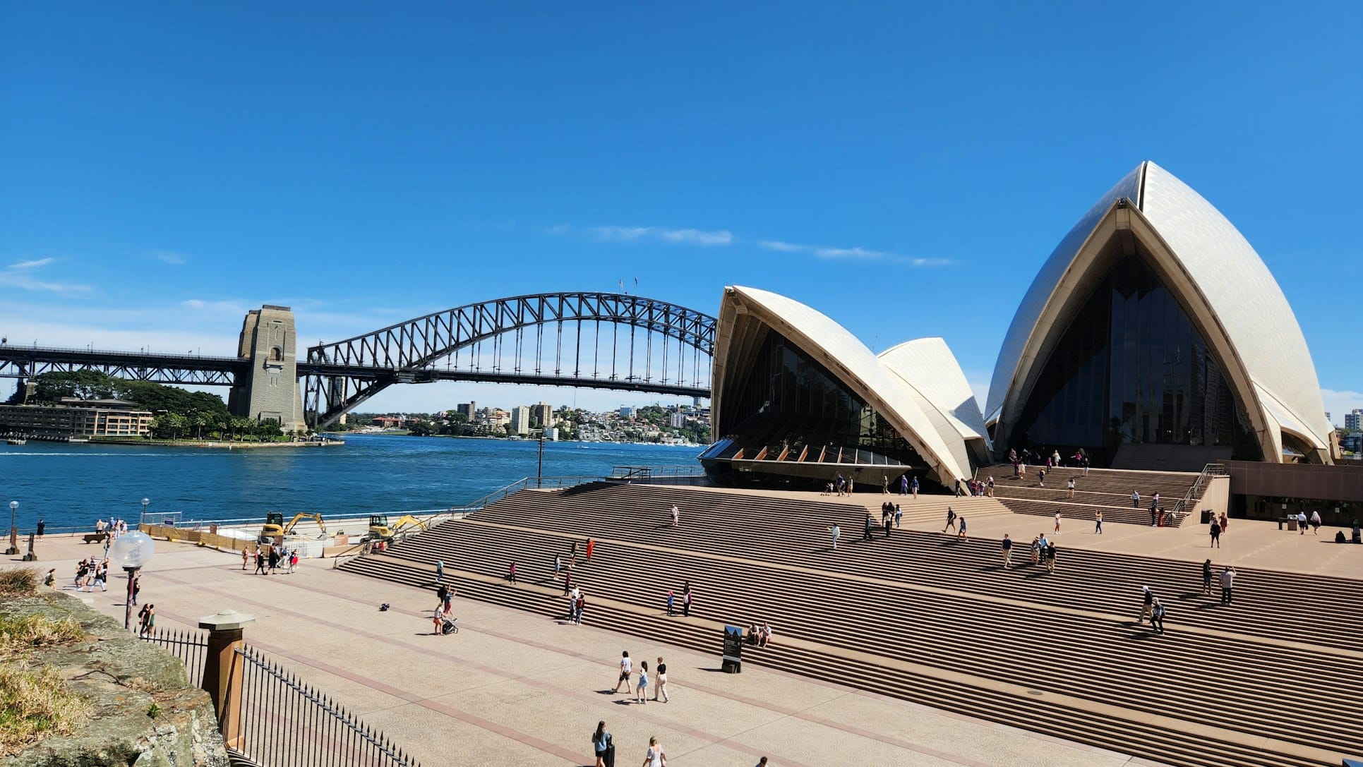 Sydney Opera House com pessoas caminhando por perto e a Sydney Harbour Bridge ao fundo, sob um céu azul claro. A foto ilustra o post de o que fazer em Sydney.