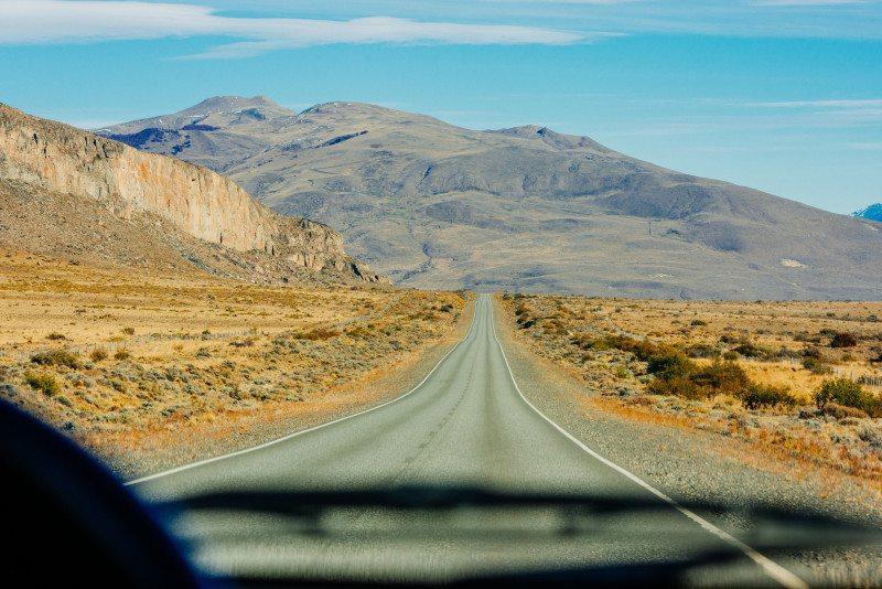 Uma estrada reta e vazia atravessa uma paisagem árida com montanhas ao fundo sob um céu azul claro.