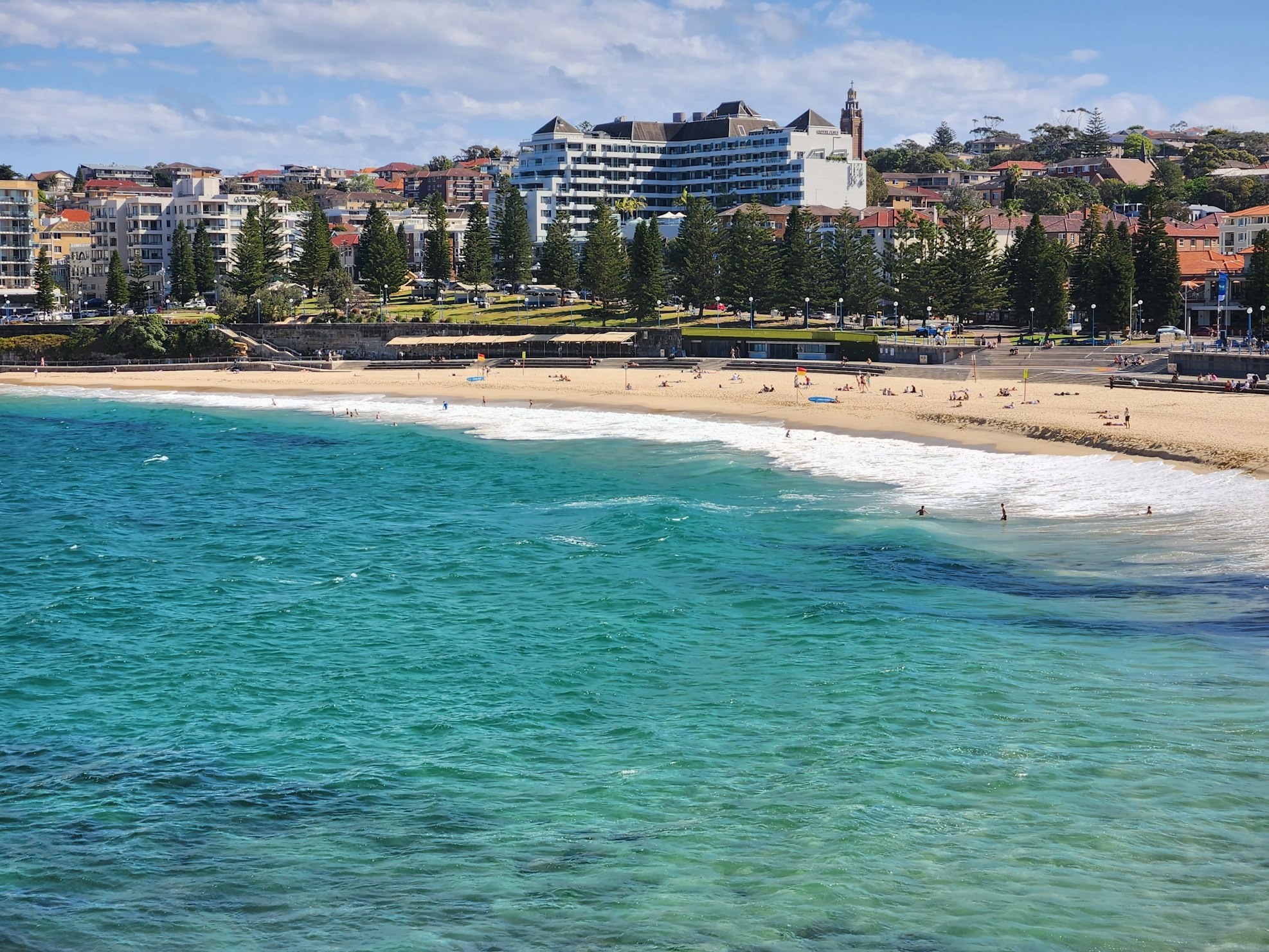 Coogee Beach com águas cristalinas azul-turquesa, pessoas tomando sol na areia, cercada por árvores e edifícios ao fundo sob um céu azul.