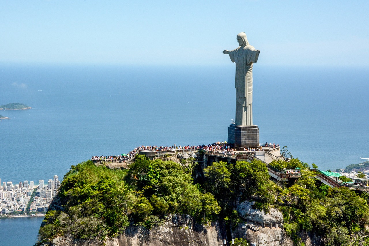 Estátua do Cristo Redentor em uma montanha com pessoas ao redor da base, com vista para a cidade e o oceano sob um céu azul claro. Essa é a paisagem de uma das trilhas no Rio de Janeiro.