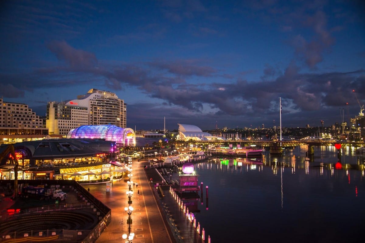 Vista noturna da Darling Harbour à beira-mar com edifícios iluminados, um caminho iluminado e barcos em águas calmas sob um céu nublado.