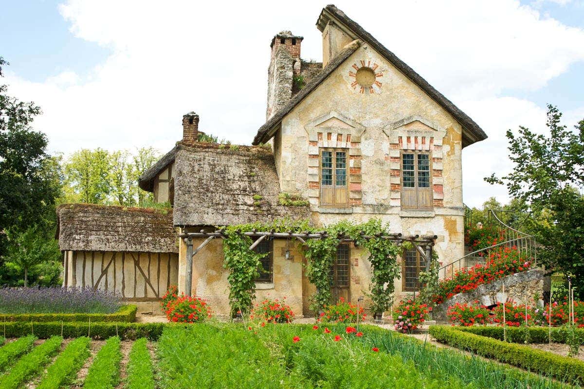 Uma casa rústica com hera, telhado de palha e um jardim de flores ao redor em um dia ensolarado. É o domínio de Maria Antonieta no Palácio de Versalhes.