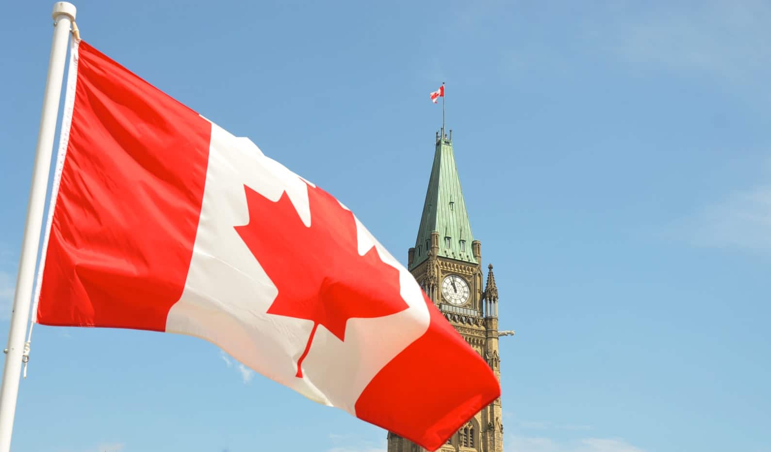 Bandeira canadense tremulando em primeiro plano com a Torre da Paz do Parlamento em Ottawa visível ao fundo.
