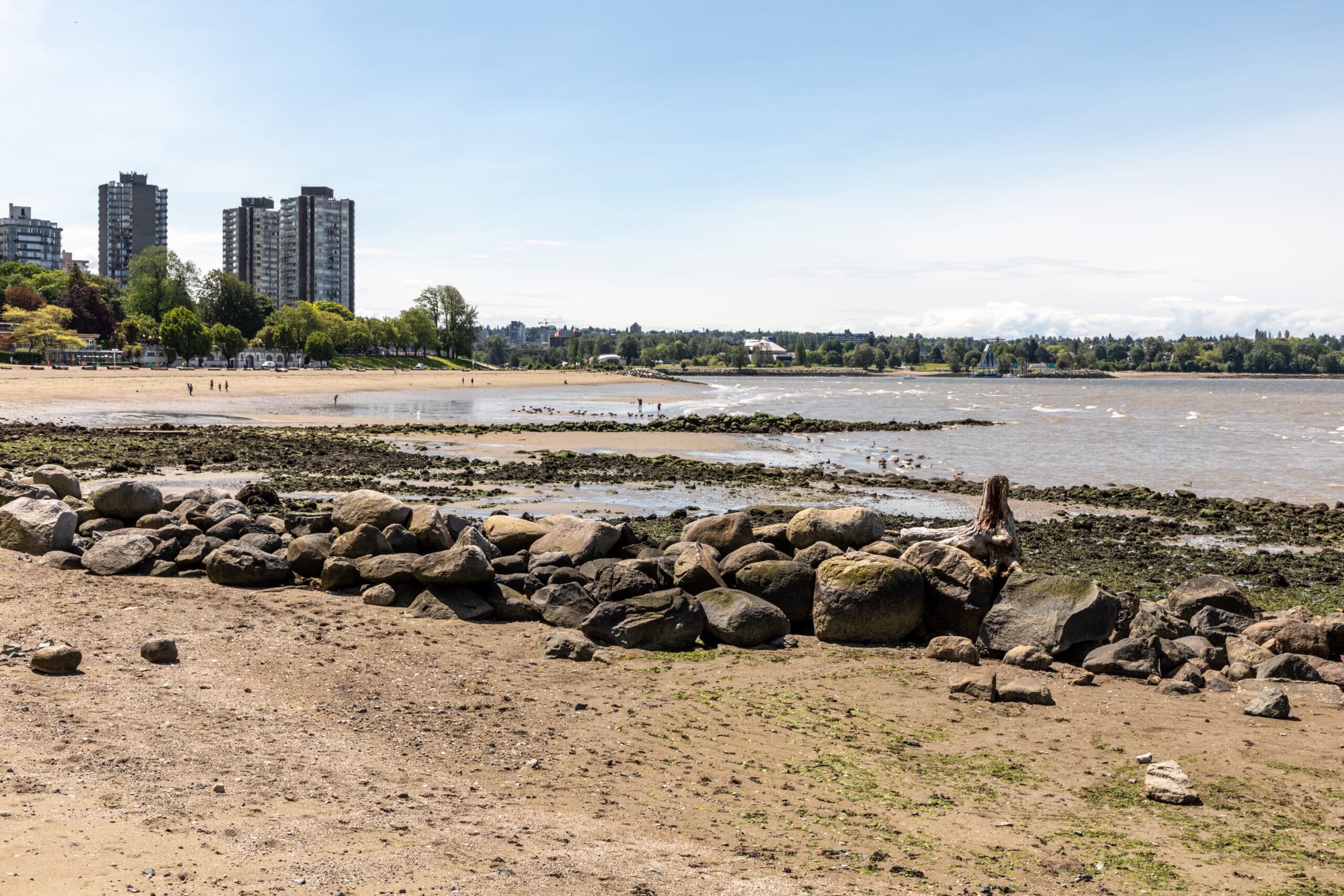 English Bay Beach com grandes pedras em primeiro plano e edifícios altos ao fundo em um dia claro.
