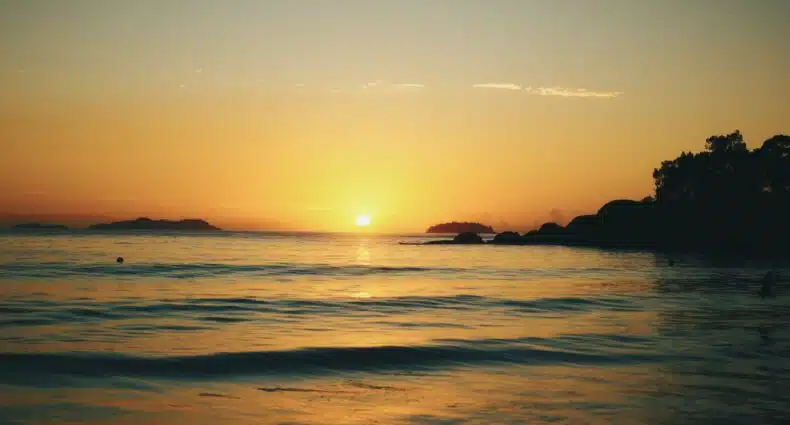Pôr do sol sobre um oceano calmo com uma silhueta de árvores e colinas no horizonte, céu com gradiente de laranja para azul. Essa é a Praia do Félix, em Ubatuba, no Litoral Norte de São Paulo.