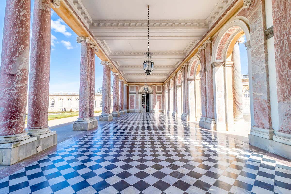 Grand Trianon no Palácio de Versalhes com um grande corredor de piso quadriculado com colunas de mármore rosa e janelas arqueadas. Uma luminária pendurada está centralizada. A luz do sol brilhante preenche o espaço.