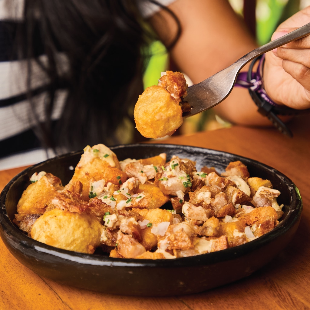 Uma pessoa segura um garfo sobre uma tigela preta cheia de comida frita, incluindo pedaços crocantes e pedaços cobertos com ervas, sobre uma mesa de madeira do Hacienda Orijen. Imagem para ilustrar post sobre roteiro em Medellín.