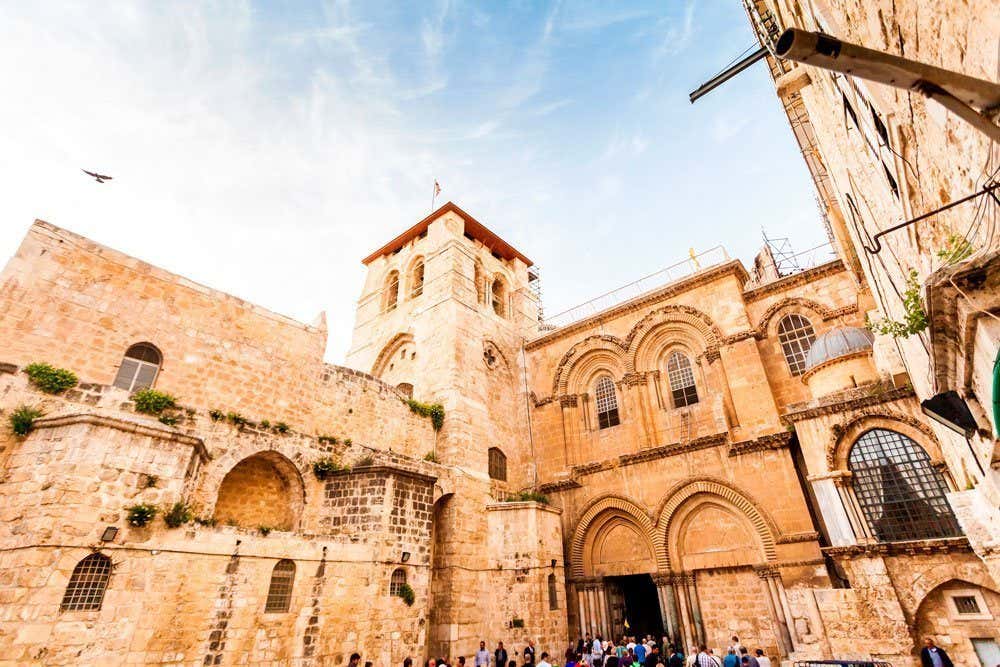 Um grupo de pessoas entra na Igreja do Santo Sepulcro, um edifício histórico de pedra com arcos e uma torre central, sob um céu azul em Jerusalém. Representa o que fazer em Israel.
