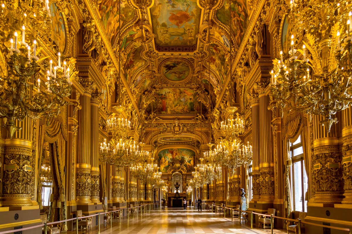 Hall ornamentado no Palácio de Versalhes com teto alto adornado com afrescos, detalhes dourados, lustres e janelas altas. Representa um grande design arquitetônico.