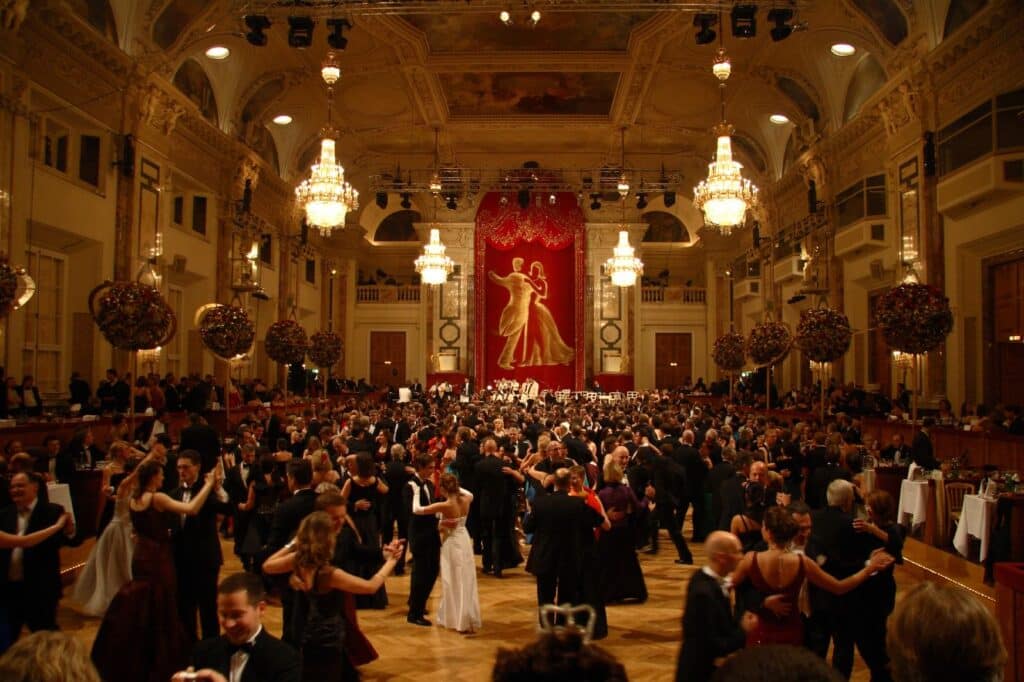 Salão do Hofburg repleto de pessoas elegantemente vestidas dançando sob lustres ornamentados e uma grande cortina vermelha ao fundo. Esse é o Kaffeesiederball, que aconteceu em 2007.