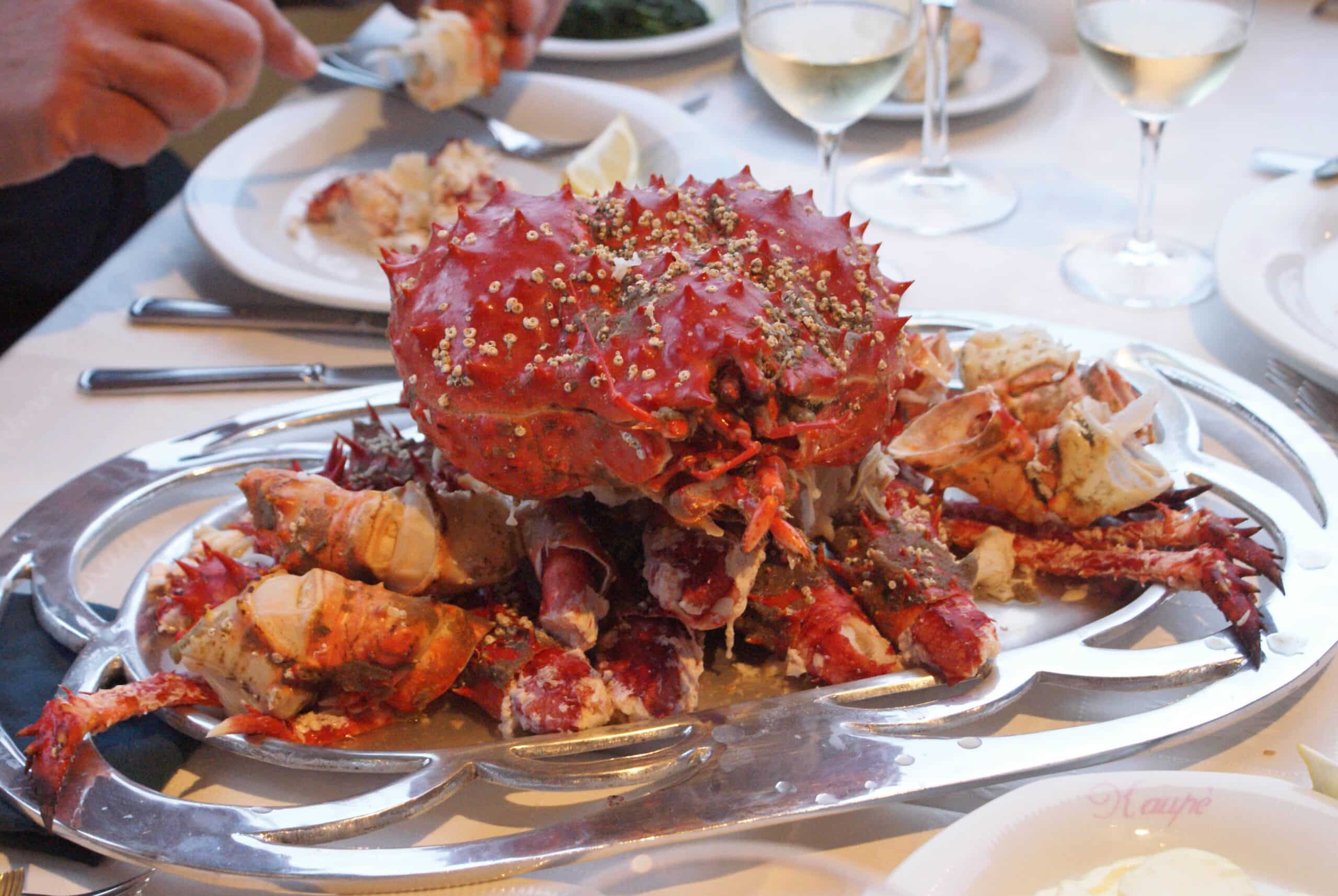 Um caranguejo cozido apresentado em uma bandeja de prata decorativa com taças de vinho e utensílios ao fundo, em uma mesa de jantar.