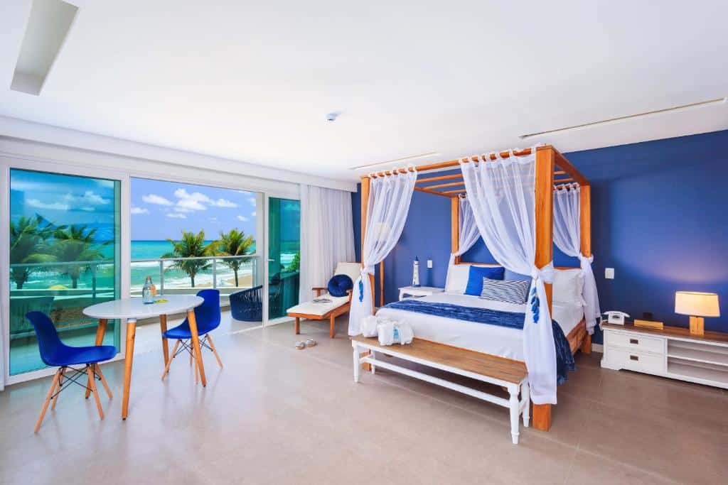 Quarto moderno do Kembali Hotel em Porto de Galinhas com cama de dossel, detalhes em azul e vista para o mar. Possui mesa redonda, cadeiras e grandes portas de correr de vidro.