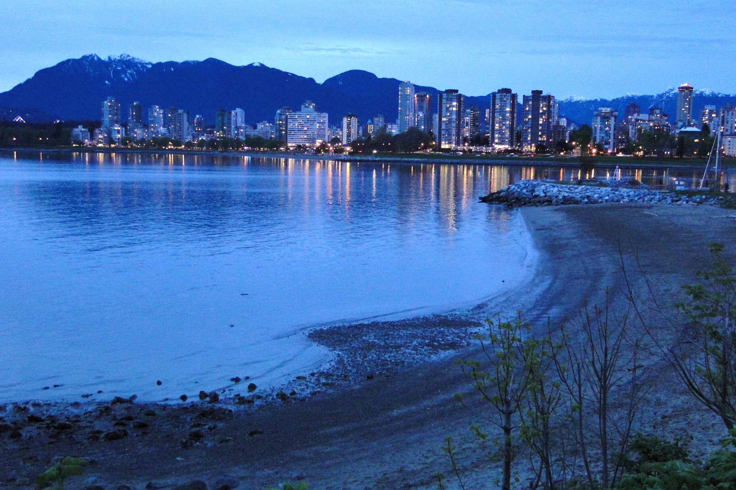 Kitsilano Beach ao anoitecer com edifícios iluminados refletindo em águas calmas, montanhas ao fundo e uma praia de areia em primeiro plano.