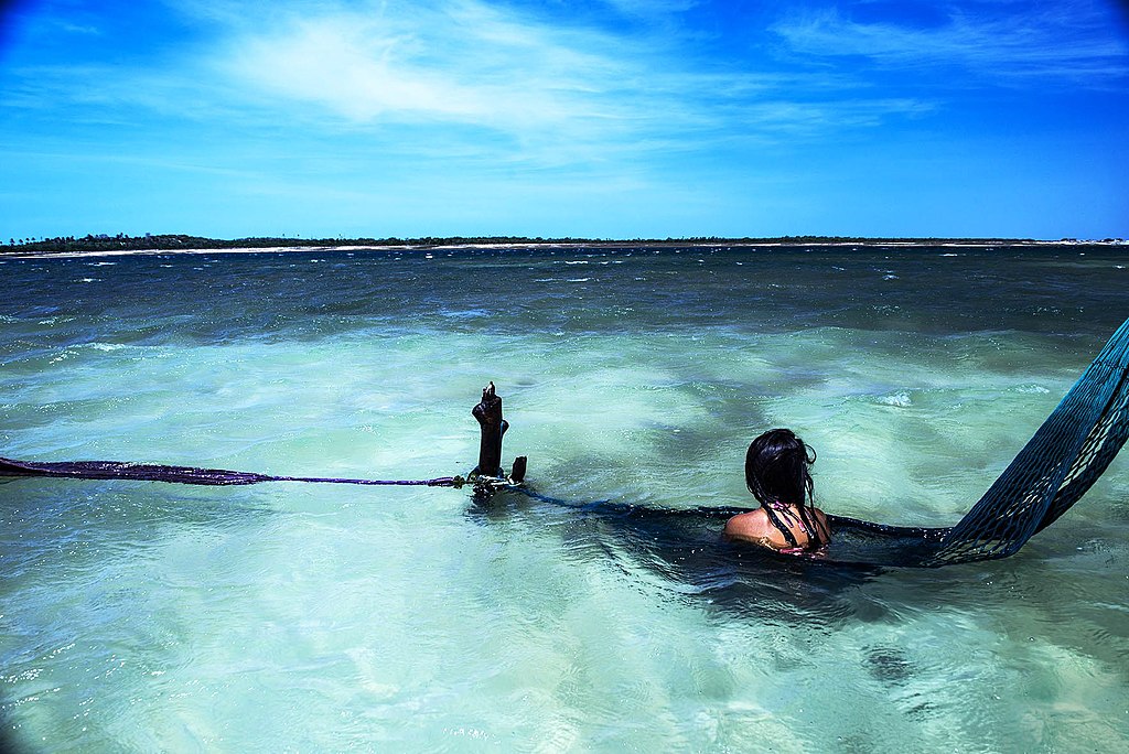 Uma pessoa está deitada em águas rasas do oceano com uma rede amarrada aos pés, sob um céu azul brilhante perto de uma costa distante.