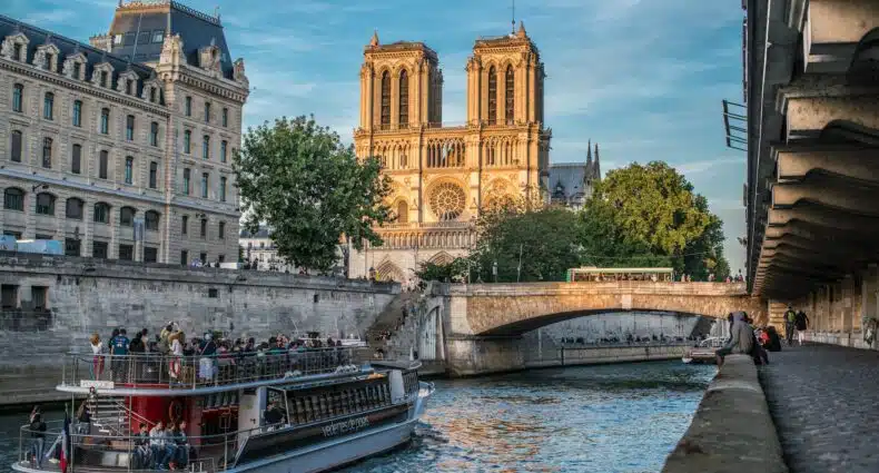 Um barco navega no Rio Sena perto da Catedral de Notre-Dame, em Paris, com edifícios históricos e uma ponte ao fundo. A imagem ilustra o post sobre quando ir para Paris.
