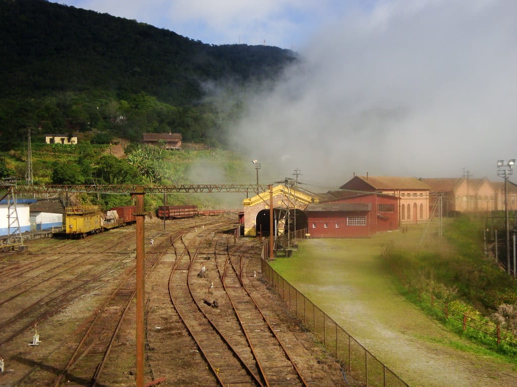 Trilhos de trem levam a um prédio amarelo e vermelho perto de uma colina florestada. A neblina obscurece parcialmente o céu. Vários vagões são visíveis.