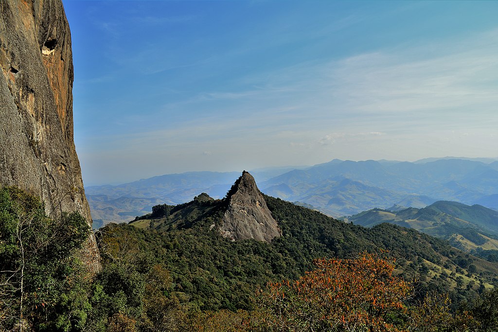 Um pico rochoso cercado por colinas verdejantes e arborizadas sob um céu azul claro, com montanhas distantes no horizonte.
