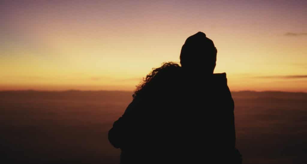 Silhueta de casal se abraçando enquanto assistem ao pôr do sol. O céu exibe tons de laranja e roxo, com uma linha do horizonte à distância.