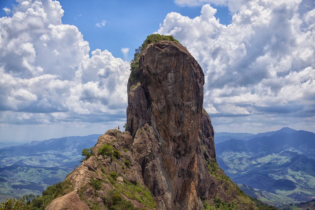 Uma formação rochosa imponente com vegetação verde sob um céu nublado. Duas pessoas estão perto da base, com vista para uma vasta paisagem. Essa é a Pedra do Baú acessada por uma das trilhas em SP.
