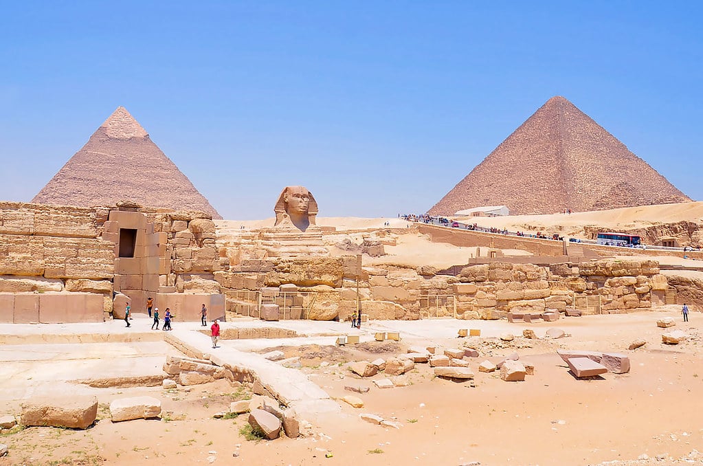 A Grande Esfinge e duas pirâmides sob um céu azul claro, com turistas caminhando perto das estruturas antigas no deserto.