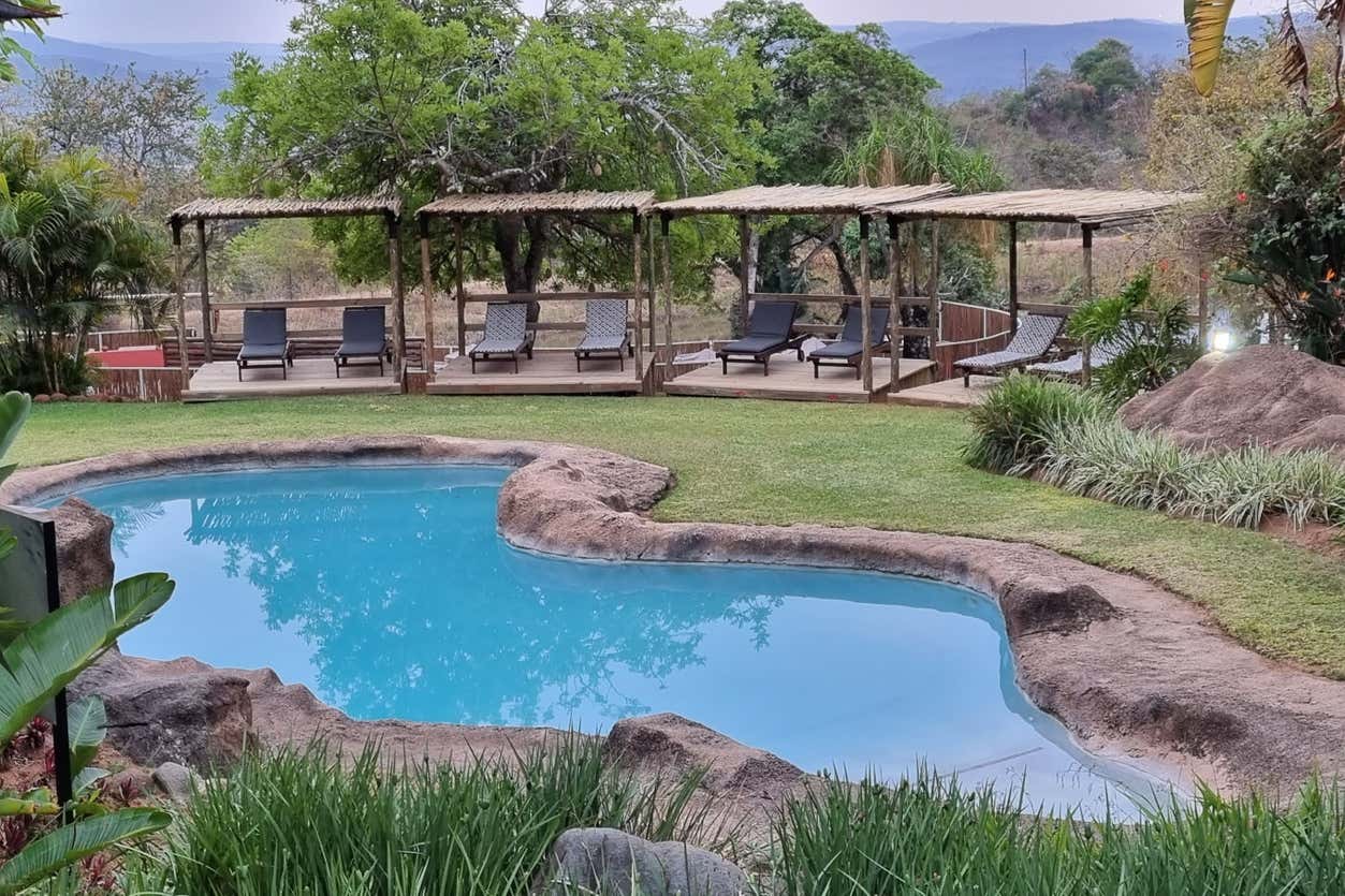 Uma piscina de estilo natural com bordas rochosas em um lodge no Kruger National Park, é cercada por grama, árvores e áreas de estar sob marquises em um ambiente de jardim.