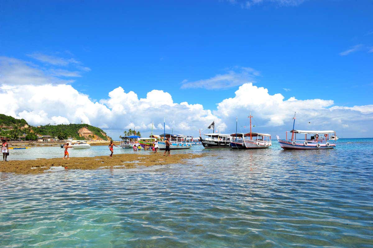 Vários barcos estão ancorados em águas costeiras claras e rasas. Pessoas caminham em uma área rochosa. Nuvens e céu azul estão ao fundo.