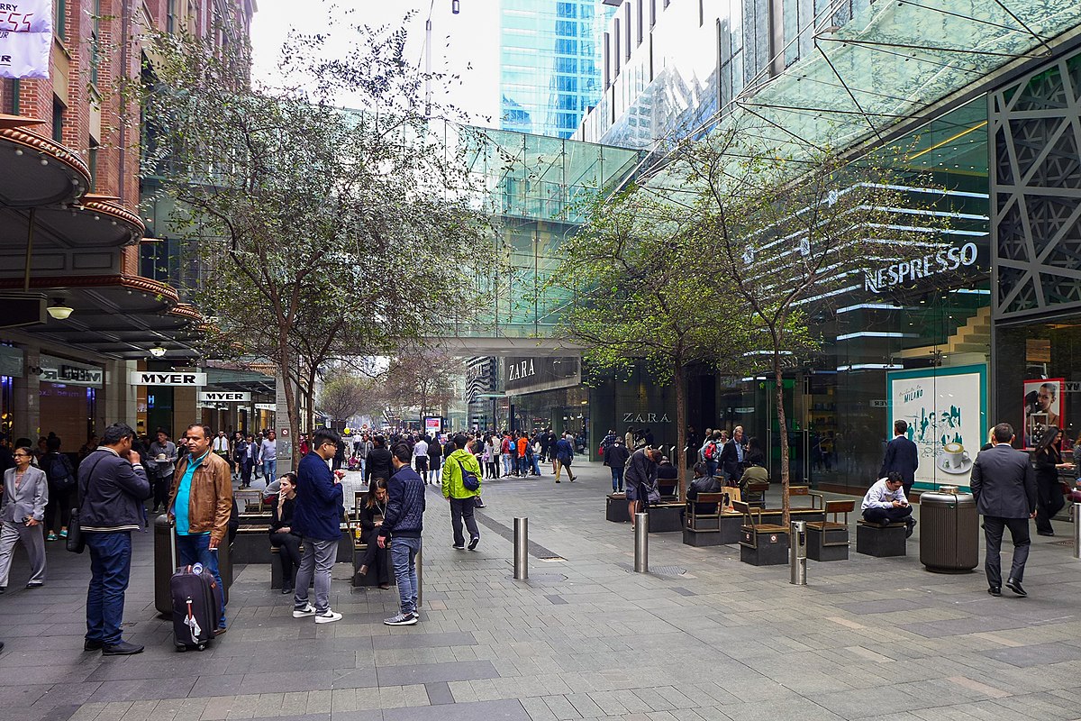 Pitt Street Mall movimentada. A área de compras é ao ar livre com pessoas caminhando e sentadas. As lojas incluem Nespresso, Zara e Myer. Árvores margeiam a rua.