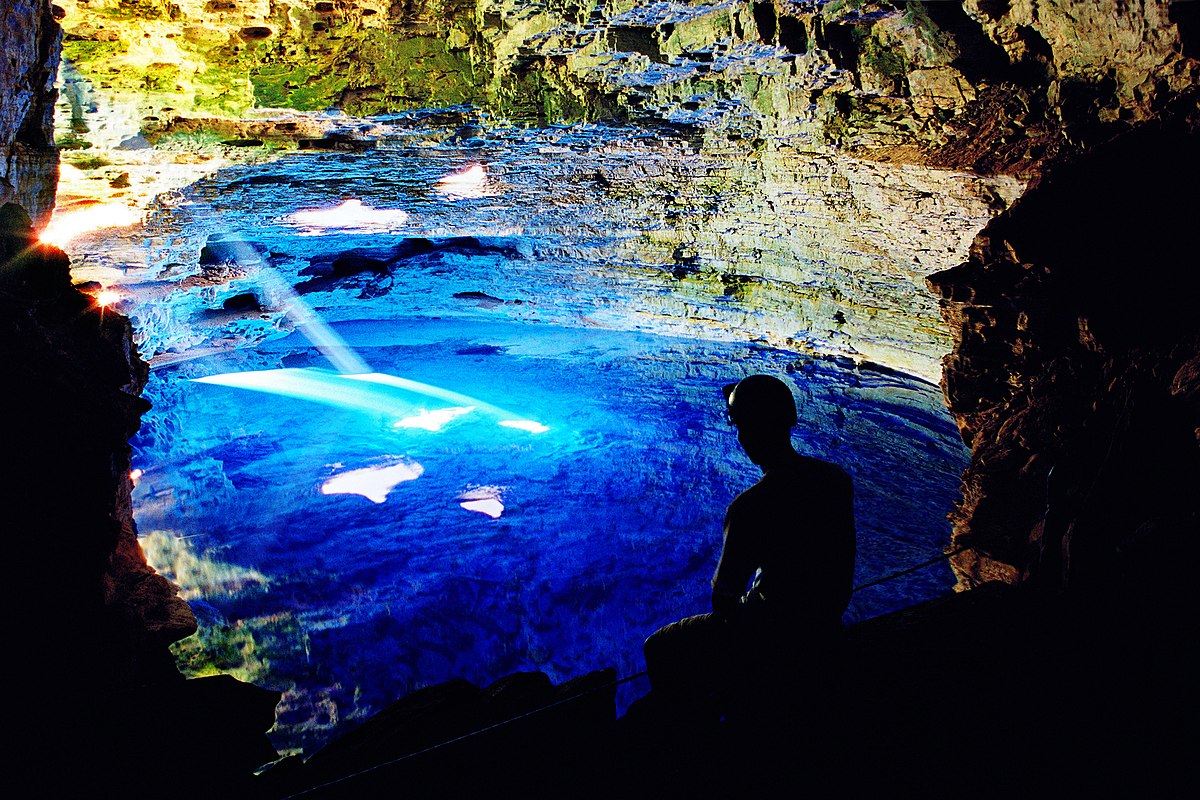 Uma pessoa senta-se perto do Poço Encantado bem iluminado com água azul. A luz do sol entra, iluminando as paredes rochosas. A foto ilustra o post de o que fazer na Chapada Diamantina.